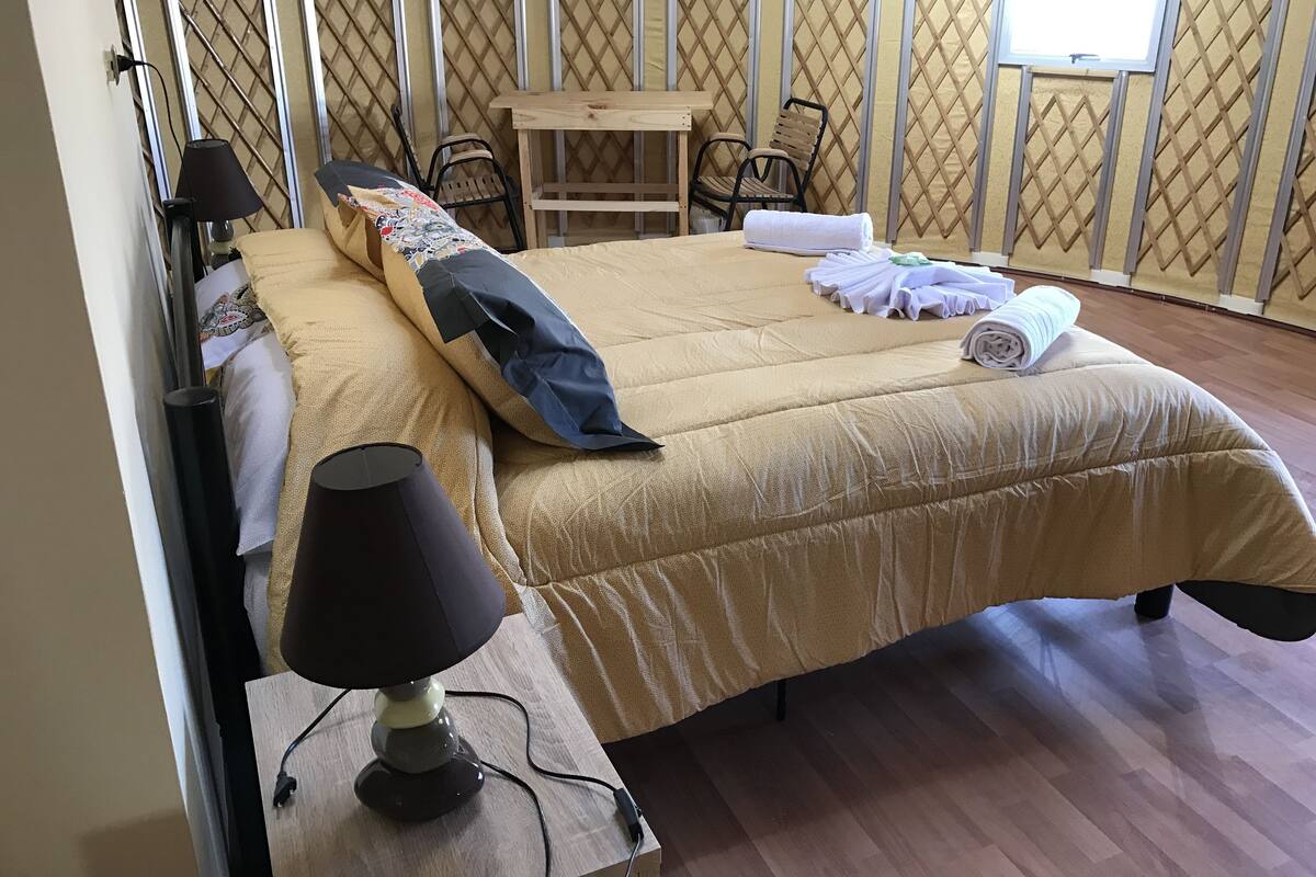 Un lit bien fait, recouvert d’une couette beige, avec un oreiller noir et une serviette blanche pliée posée dessus. On y trouve une table en bois avec deux chaises, une lampe sur une table de chevet et une fenêtre donnant sur une clôture.