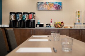 Zona de recepción de hotel con estación de café, frutero y un cuadro en la pared.