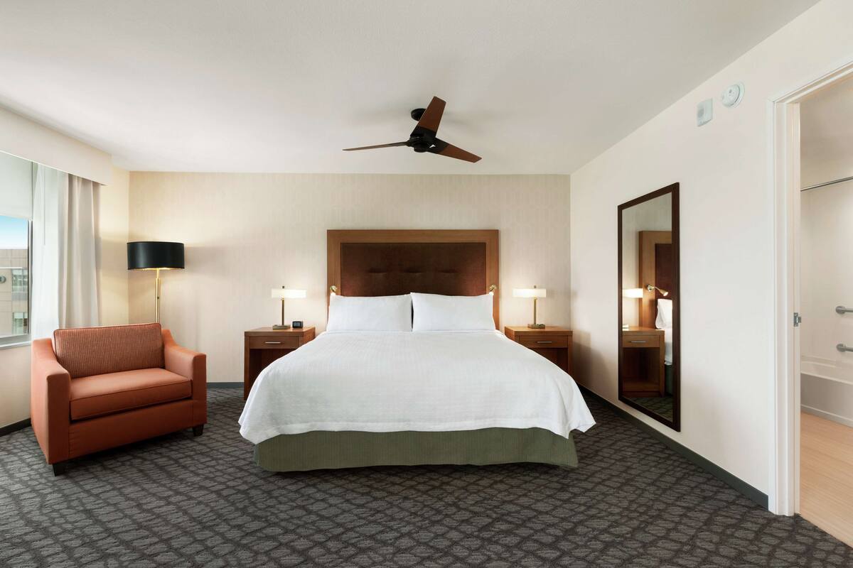 Habitación de hotel con una cama, una silla, un ventilador de techo, dos mesitas de noche con lámparas, un espejo grande y una puerta.