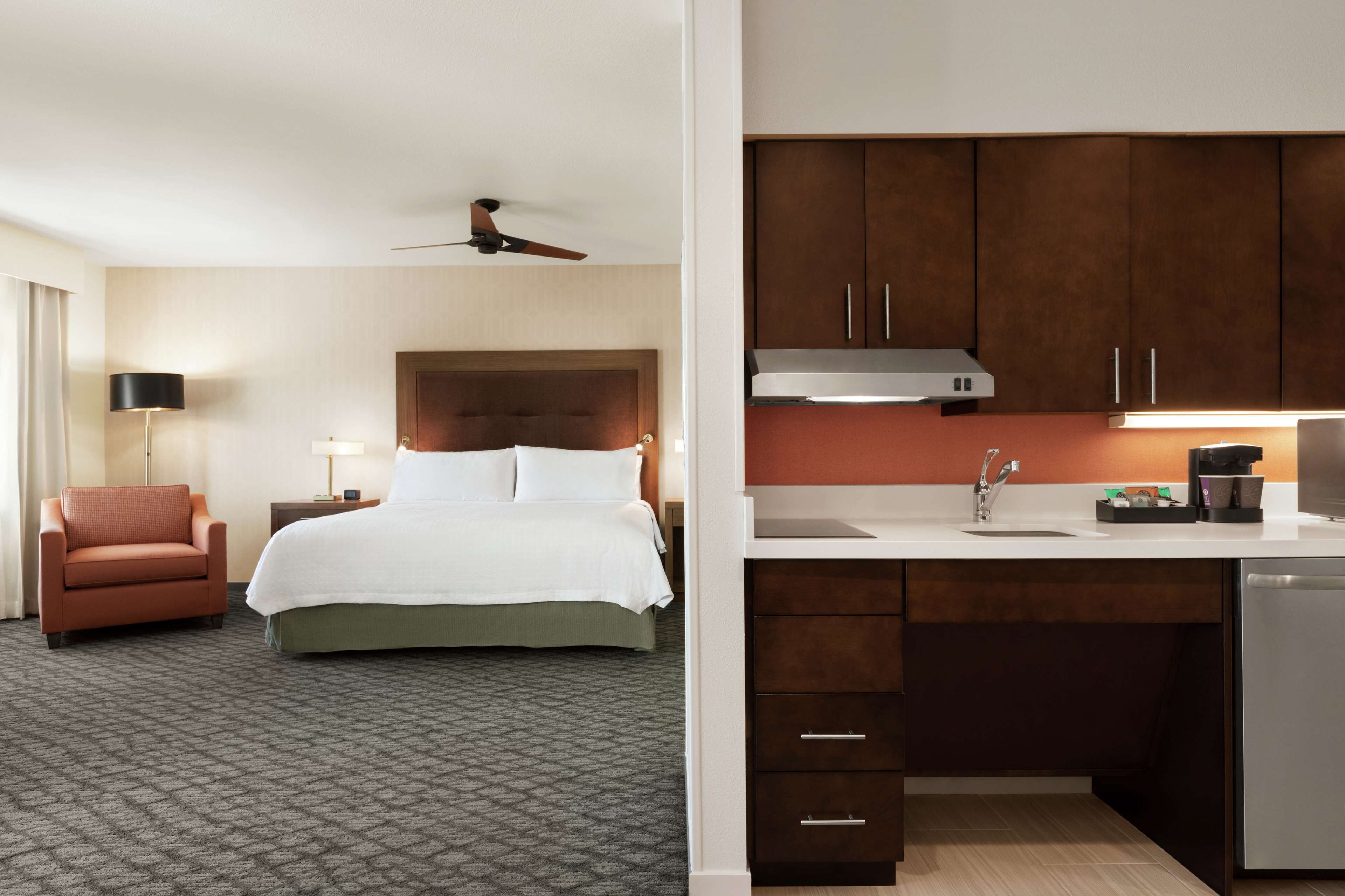 Une chambre d’hôtel avec un lit, une chaise, un bureau avec un lavabo et un ventilateur de plafond.