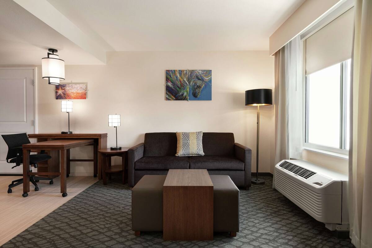 Una habitación de hotel moderna con sofá, escritorio, mesa de centro y un ventanal grande.