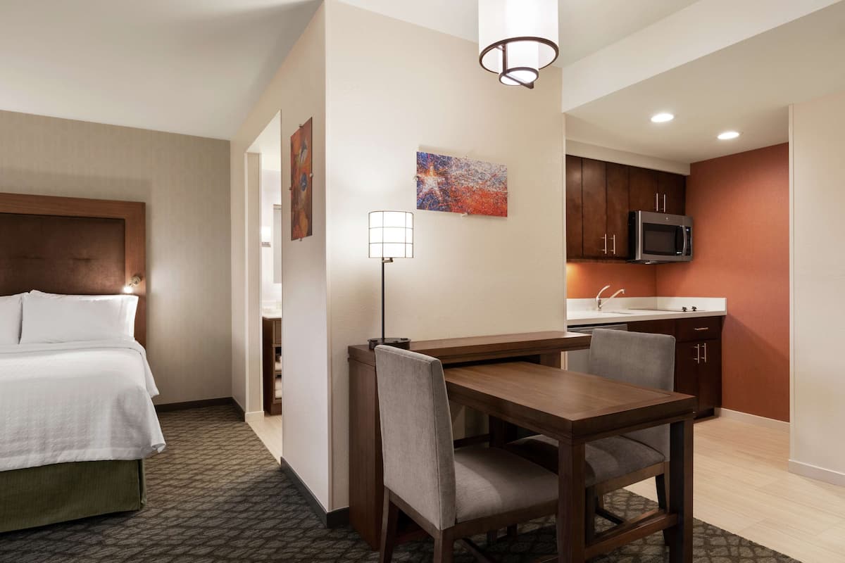 Una habitación de hotel con una cama, un escritorio con dos sillas, una lámpara, un microondas y una kitchenette pequeña.