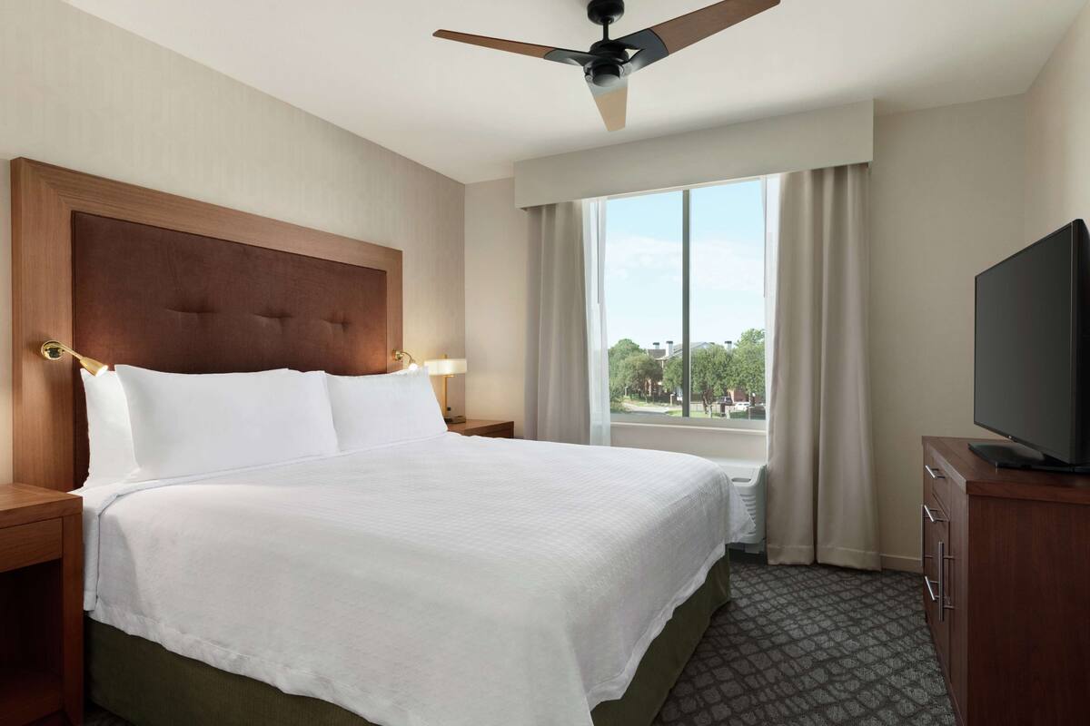 Una habitación de hotel con una cama grande, un ventilador de techo, un televisor y una ventana con cortinas.