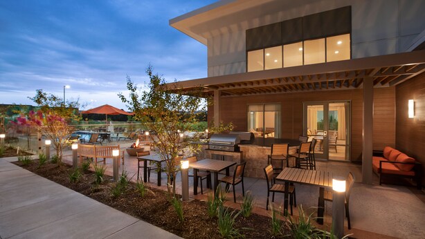 Un patio moderno al aire libre con zona de barbacoa, mobiliario y vista a una piscina al fondo.