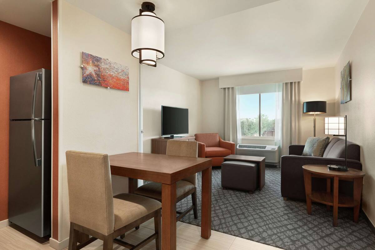 Habitación de hotel con comedor, sala de estar con sofá y sillón, televisor de pantalla plana y refrigerador.