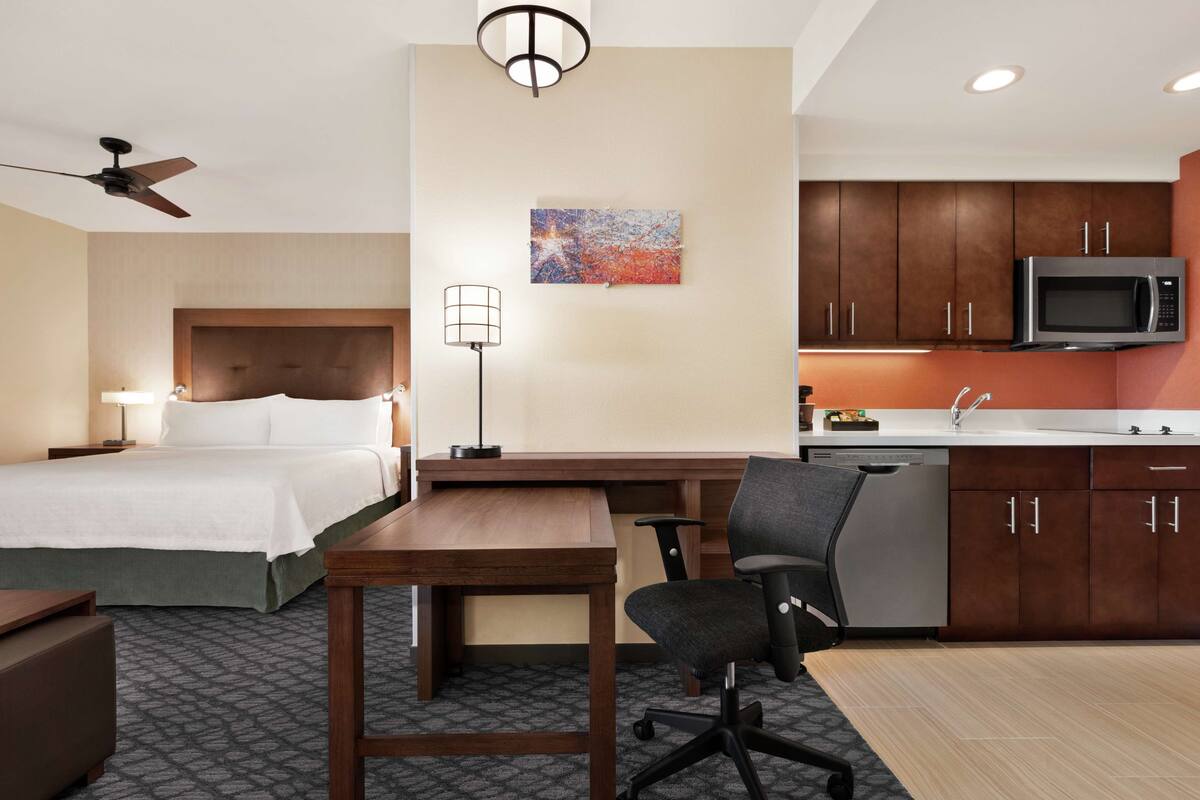 Habitación de hotel con cama, escritorio, silla, lámpara, ventilador de techo, obras de arte y zona de cocina.