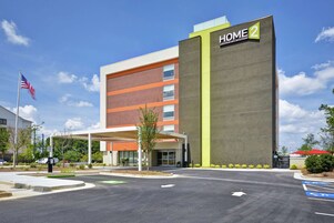 Un hotel moderno con un llamativo letrero que dice "HOME 2", un acceso cubierto y un estacionamiento con espacios para discapacitados.