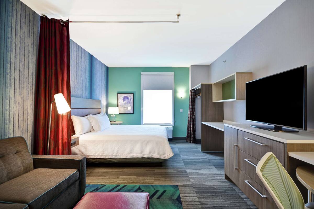 A hotel room with a bed, a desk with a flat-screen TV, a chair, a sofa, and a window with curtains.