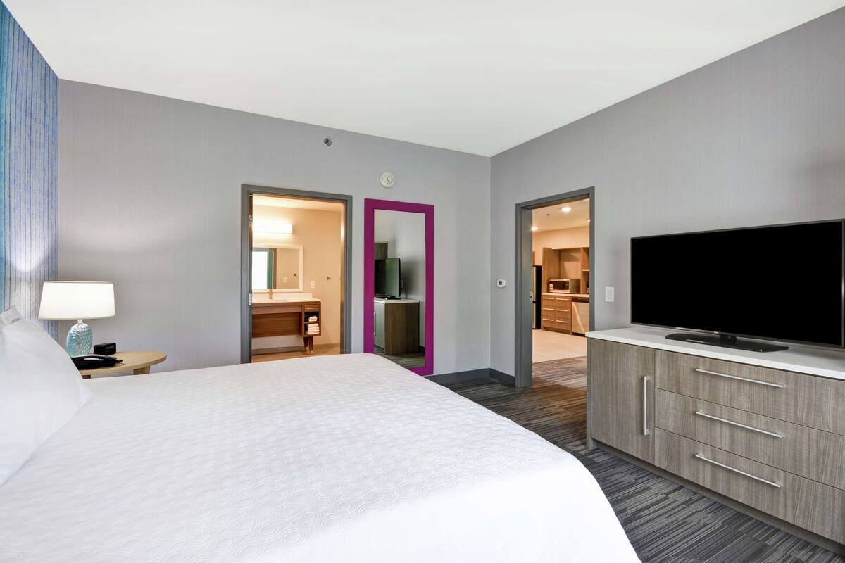 A hotel room with a large bed, a TV, a desk with a lamp, and a kitchenette visible through an open door.
