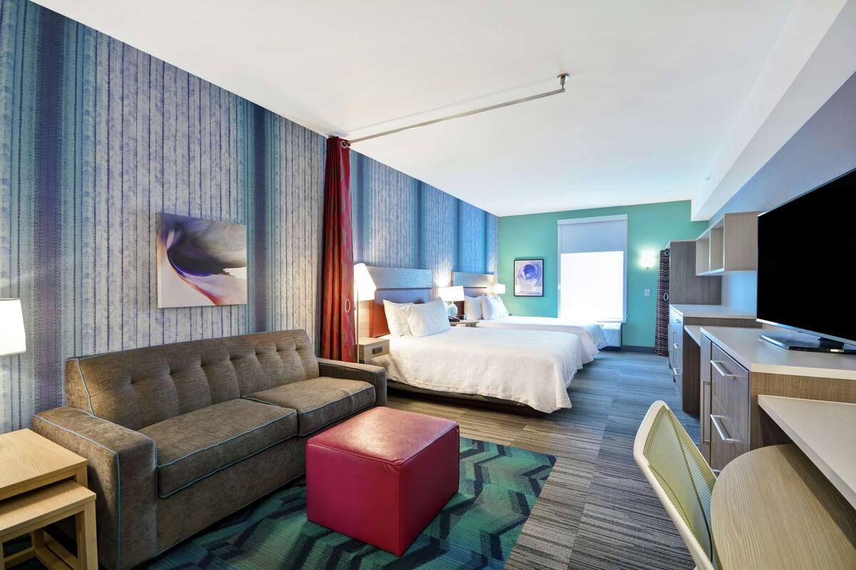 A hotel room with a sofa, a red ottoman, a bed, a desk, and a TV.