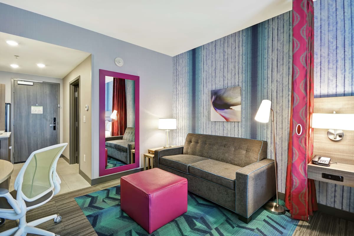A hotel room with a sofa, ottoman, and a desk with a lamp.