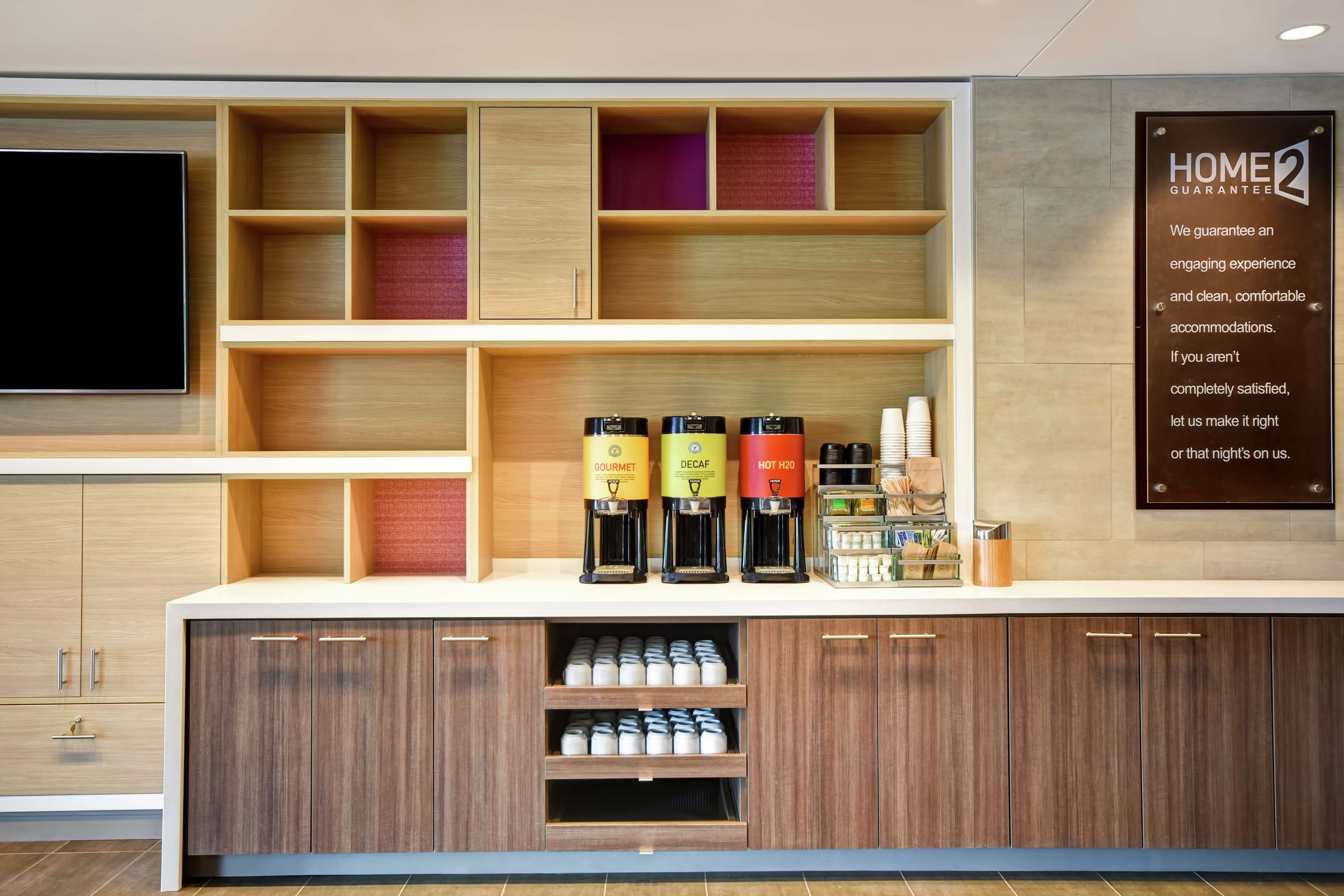 A hotel reception area with a wooden counter, multiple coffee machines, and a sign stating "HOME2 GUARANTEE".