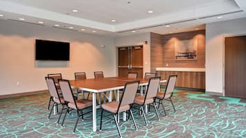 Una sala de conferencias con una mesa grande y sillas, un televisor de pantalla plana y una zona de cocina con mostrador y armarios de madera.