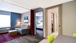 A hotel room with a sofa, a small table, a patterned carpet, and a view of a bedroom with a bed and a nightstand. - Home2 Suites by Hilton Walpole Foxboro (Walpole)