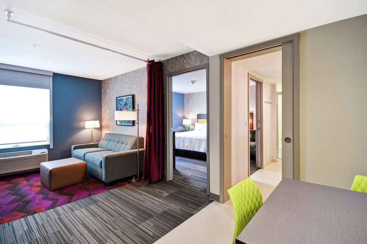 A hotel room with a sofa, a small table, a patterned carpet, and a view of a bedroom with a bed and a nightstand.