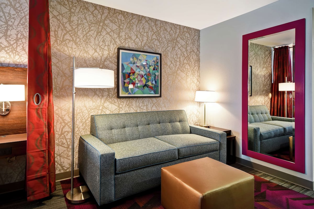 A modern hotel room with a grey sofa, a large mirror, and a framed abstract painting on the wall.