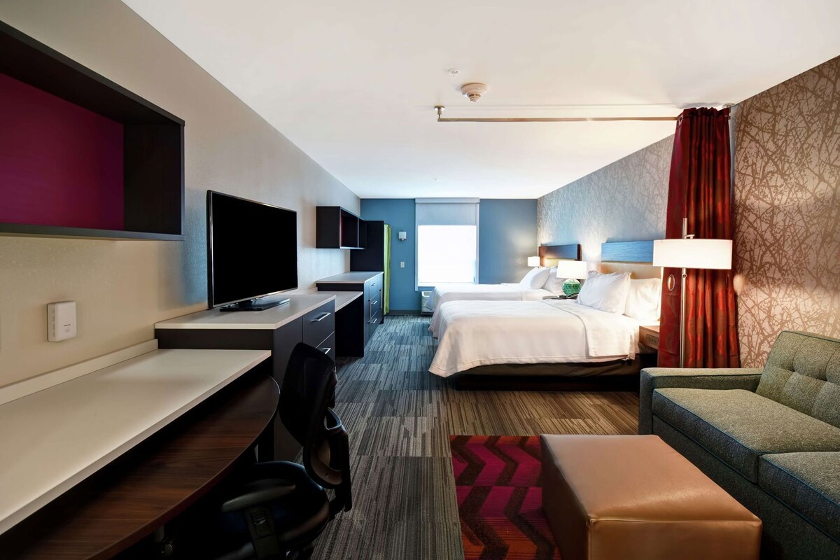 Habitación de hotel con escritorio, dos camas, televisor, sofá y alfombra estampada.