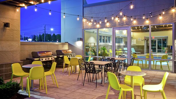 An outdoor patio with yellow chairs and tables, a barbecue grill, and string lights. - Home2 Suites by Hilton Walpole Foxboro (Walpole)