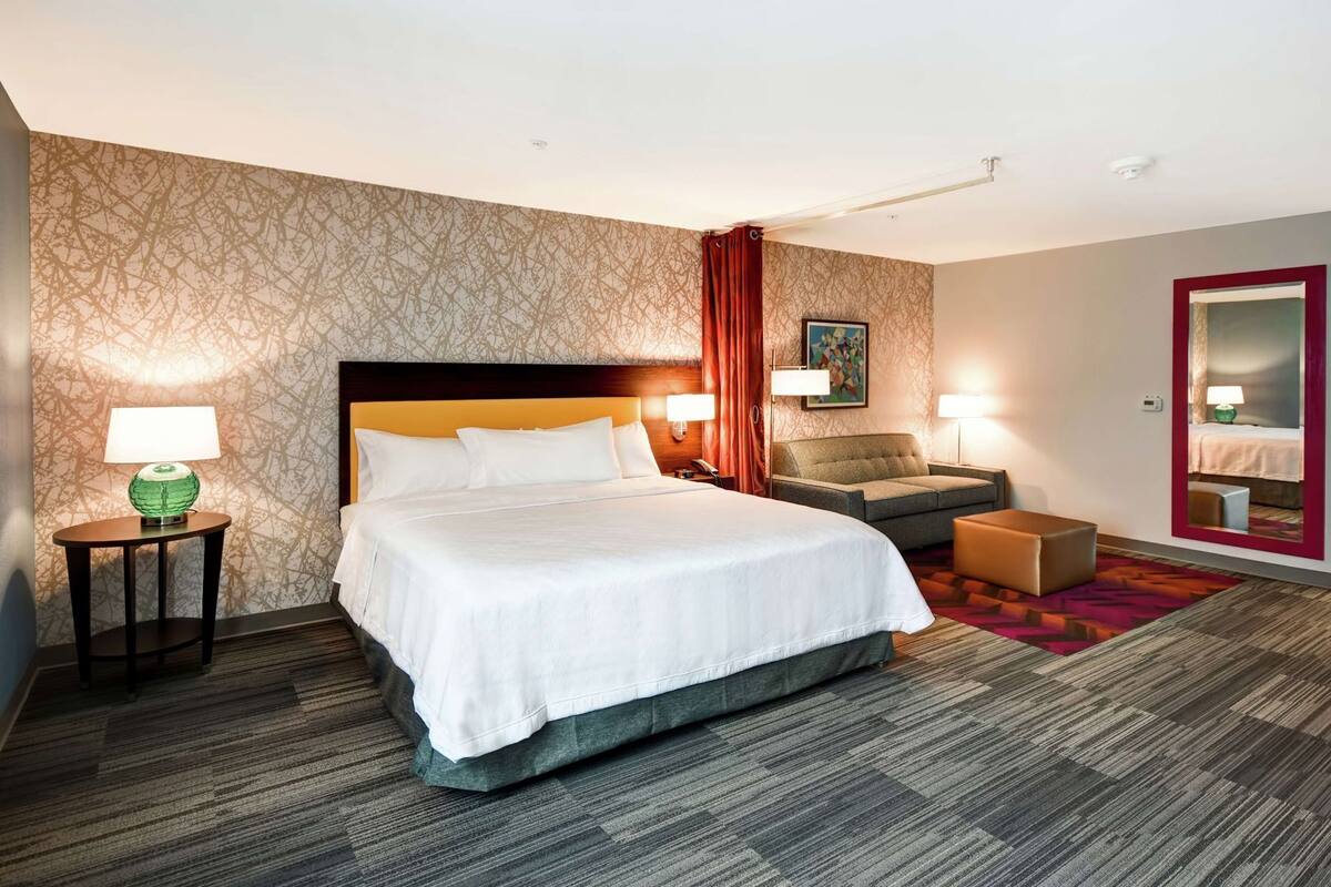 Quarto de hotel com uma cama grande, um sofá, uma mesa pequena com abajur e um espelho.