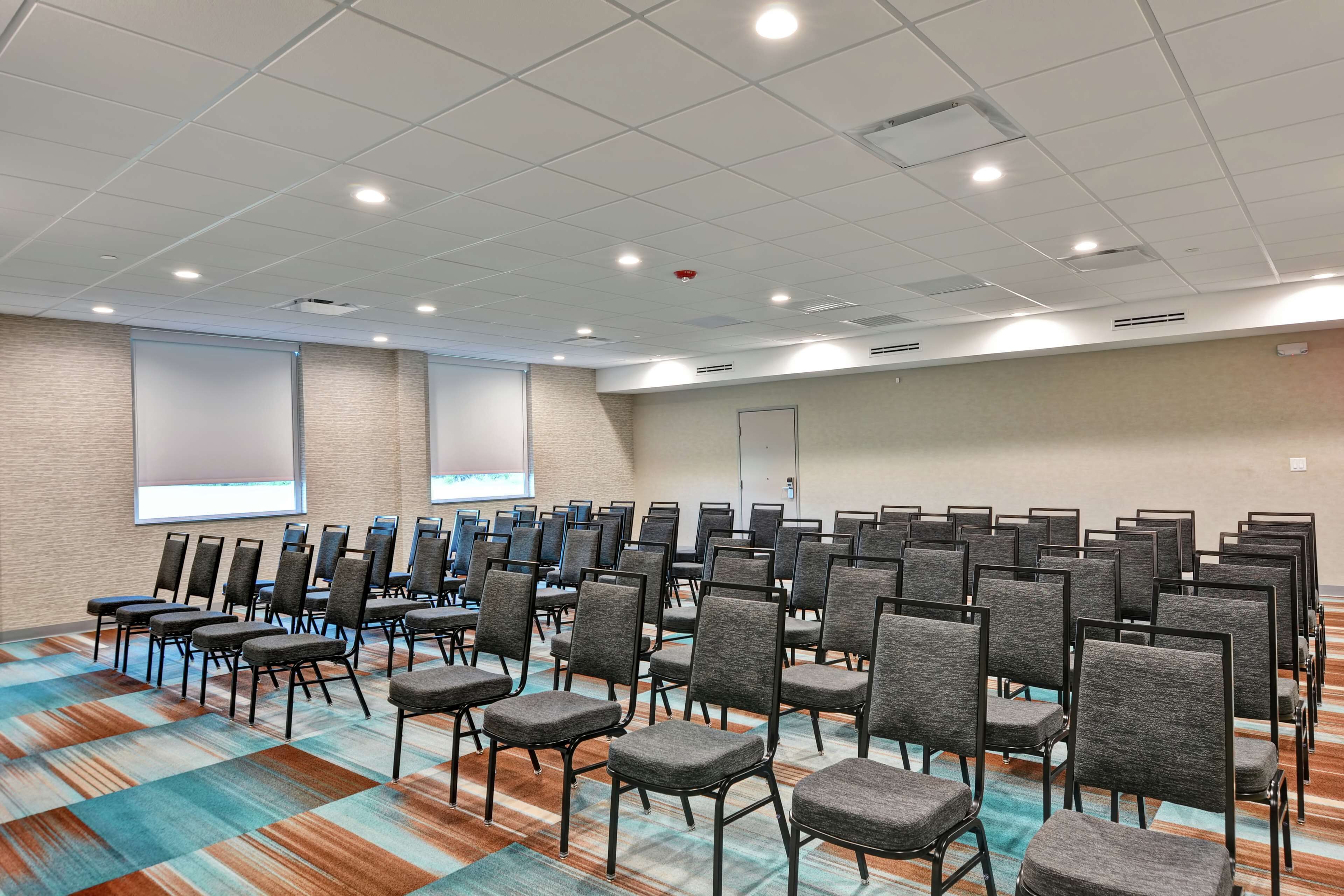 Une salle de conférence avec des rangées de chaises, un sol recouvert de moquette et un écran de projection fixé au mur.