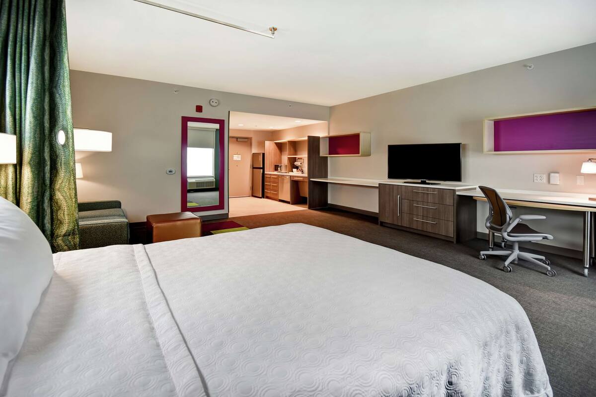 A hotel room with a large bed, a desk, a chair, a television, and a kitchenette.