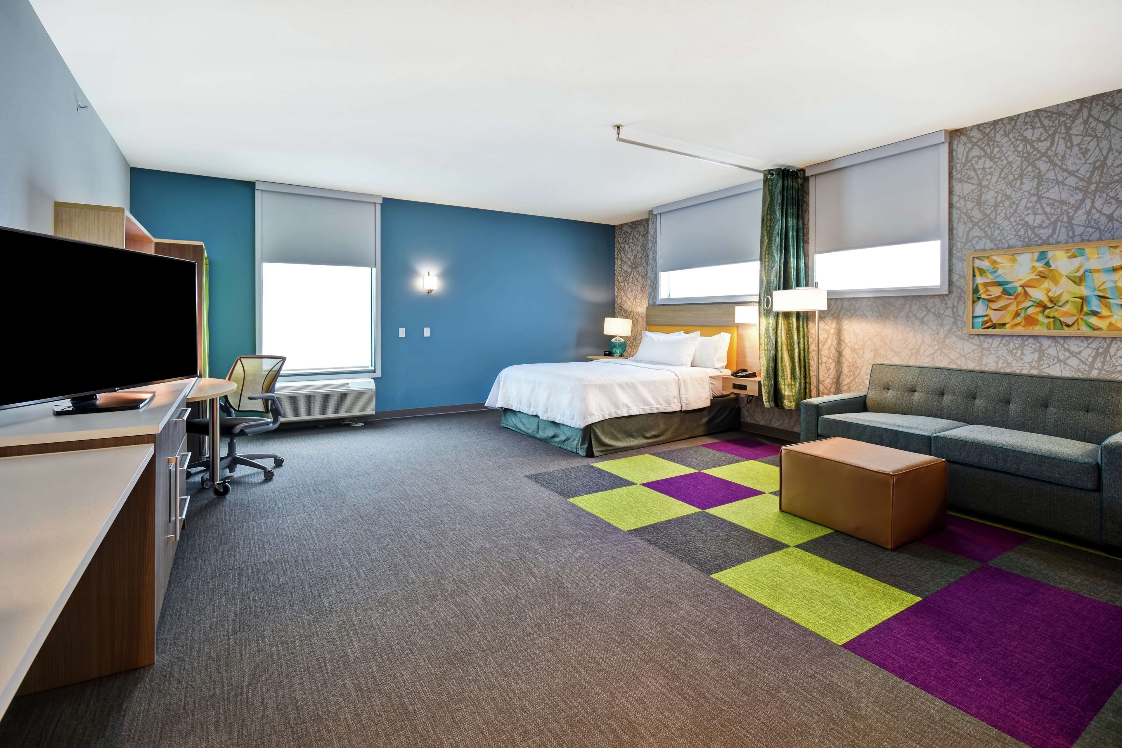 Quarto de hotel moderno com cama, sofá, escrivaninha com televisão e tapete colorido.