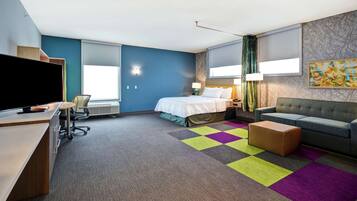 Quarto de hotel moderno com cama, sofá, escrivaninha com televisão e tapete colorido.