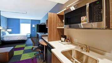 A compact hotel room with a kitchenette, a bed, a desk, and a colorful carpet.