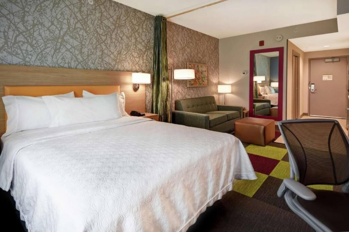A hotel room with a large bed, a green armchair, a small table, and a mirror.