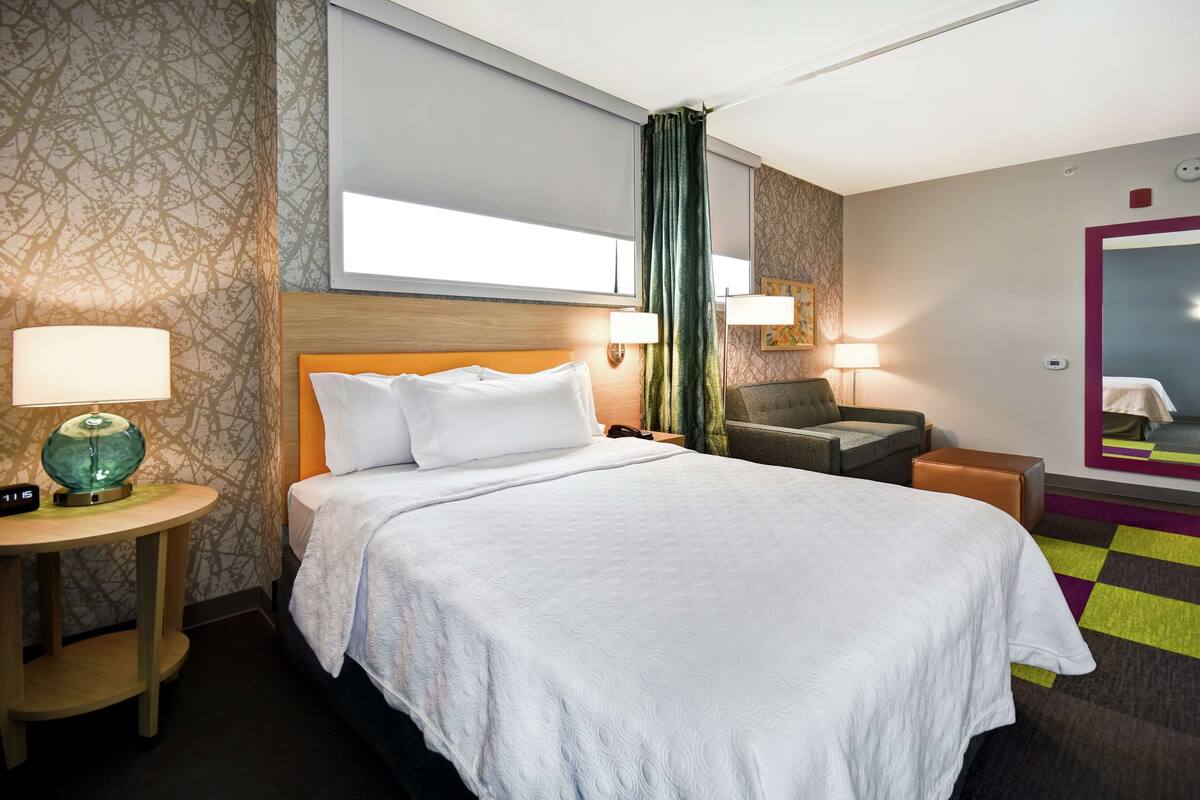 A hotel room with a large bed, a desk with a lamp, a chair, and a mirror on the wall.