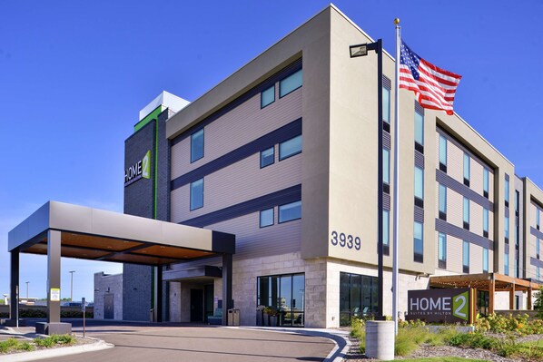 Ein modernes Hotel mit überdachter Eingangspartie, einer Flagge und dem Schriftzug "Home 2 Suites by Hilton" am Gebäude.