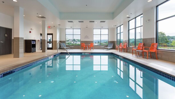 Una piscina con agua cristalina azul, rodeada de sillas anaranjadas y amplias ventanas con vistas al exterior.