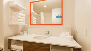 A modern bathroom with a white sink, two towel dispensers, a mirror with an orange frame, and a shelf with folded towels.
