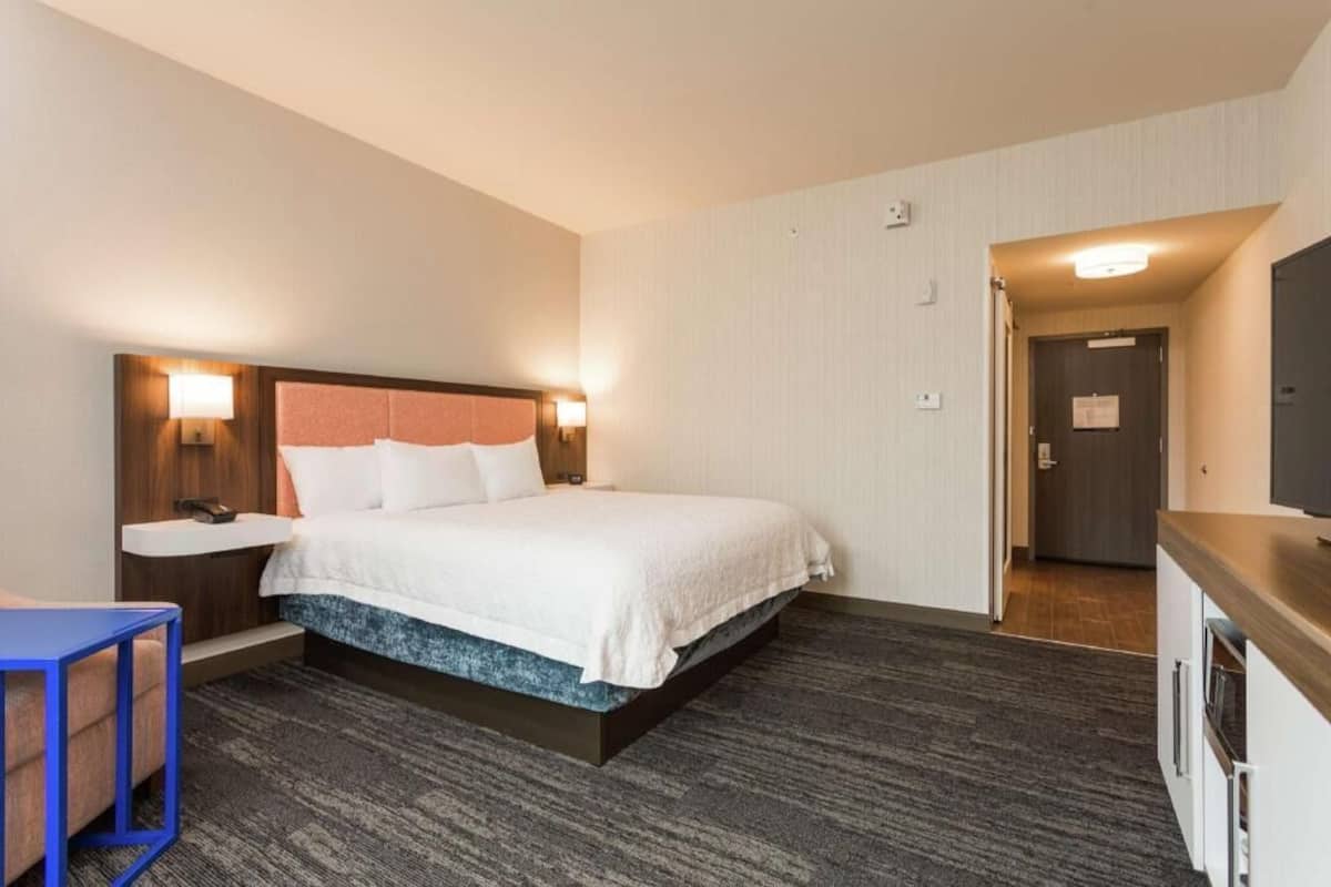 A hotel room with a bed, bedside table, lamp, and a small desk with a chair.
