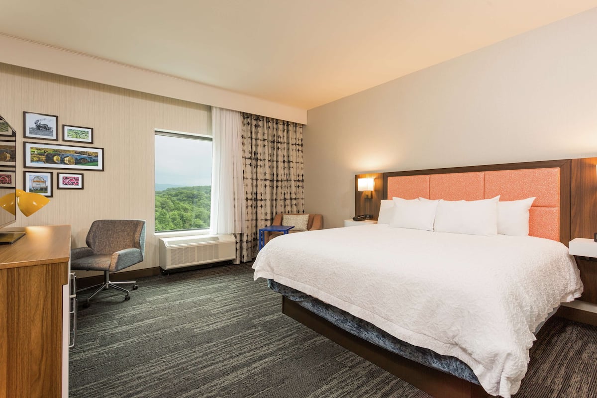 A hotel room with a large bed, a desk, a chair, a window with curtains, and framed pictures on the wall.