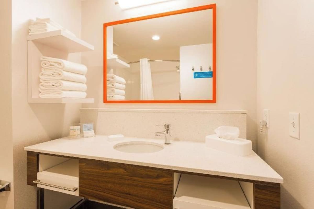 Un baño con un lavamanos blanco, un espejo con marco naranja y estantes con toallas dobladas.