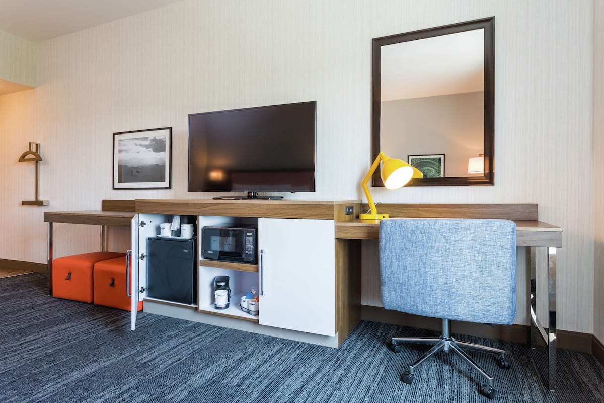 A hotel room with a desk, a television, a microwave, a coffee maker, a chair, and a mirror.