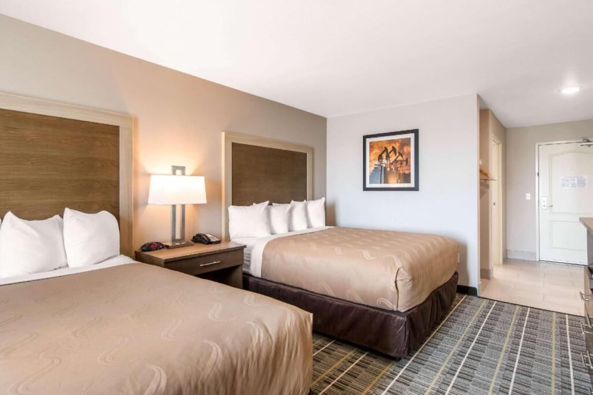 Une chambre d’hôtel avec deux lits, une table de chevet avec une lampe, un tableau encadré au mur et une porte donnant sur une autre pièce.