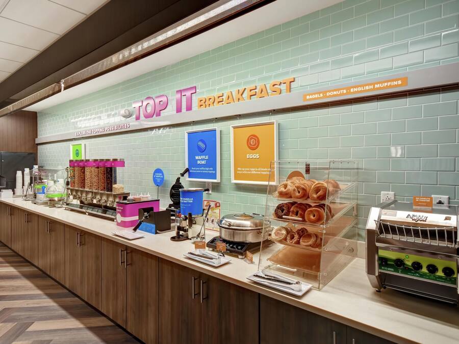 Um balcão de café da manhã com uma placa indicando 'TOP IT BREAKFAST' e opções de cardápio como waffles, ovos e muffins.