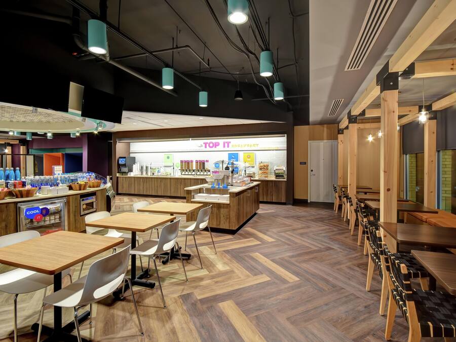 A modern cafe with wooden tables and chairs, a visible counter with a menu, and a herringbone-patterned floor.