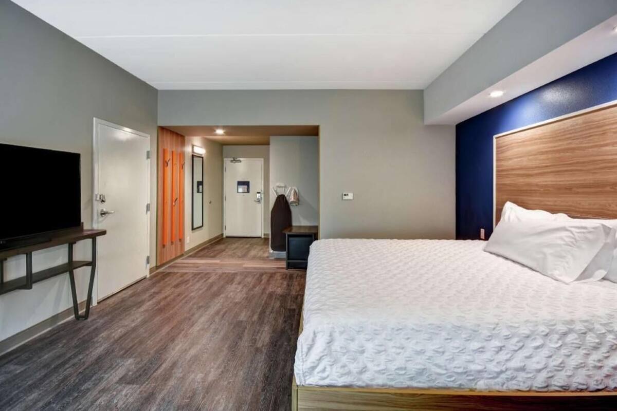 A hotel room with a bed, a TV, a wooden headboard, and a hallway leading to another room.