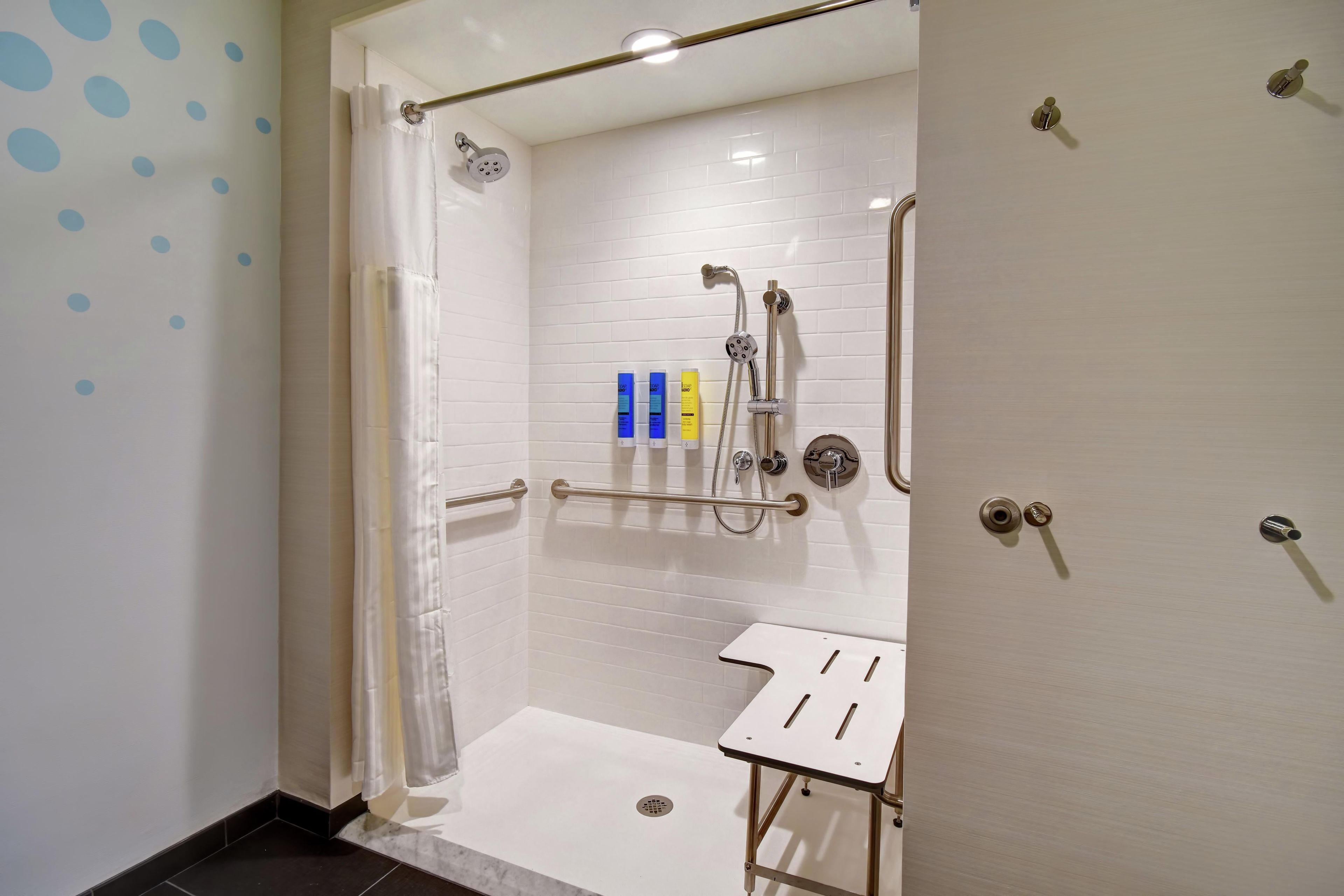 Une douche à l’italienne avec un mur recouvert de carreaux blancs, un pommeau de douche fixe, un pommeau de douche amovible, un panneau de commande de la douche et un lavabo blanc avec un cadre en métal.