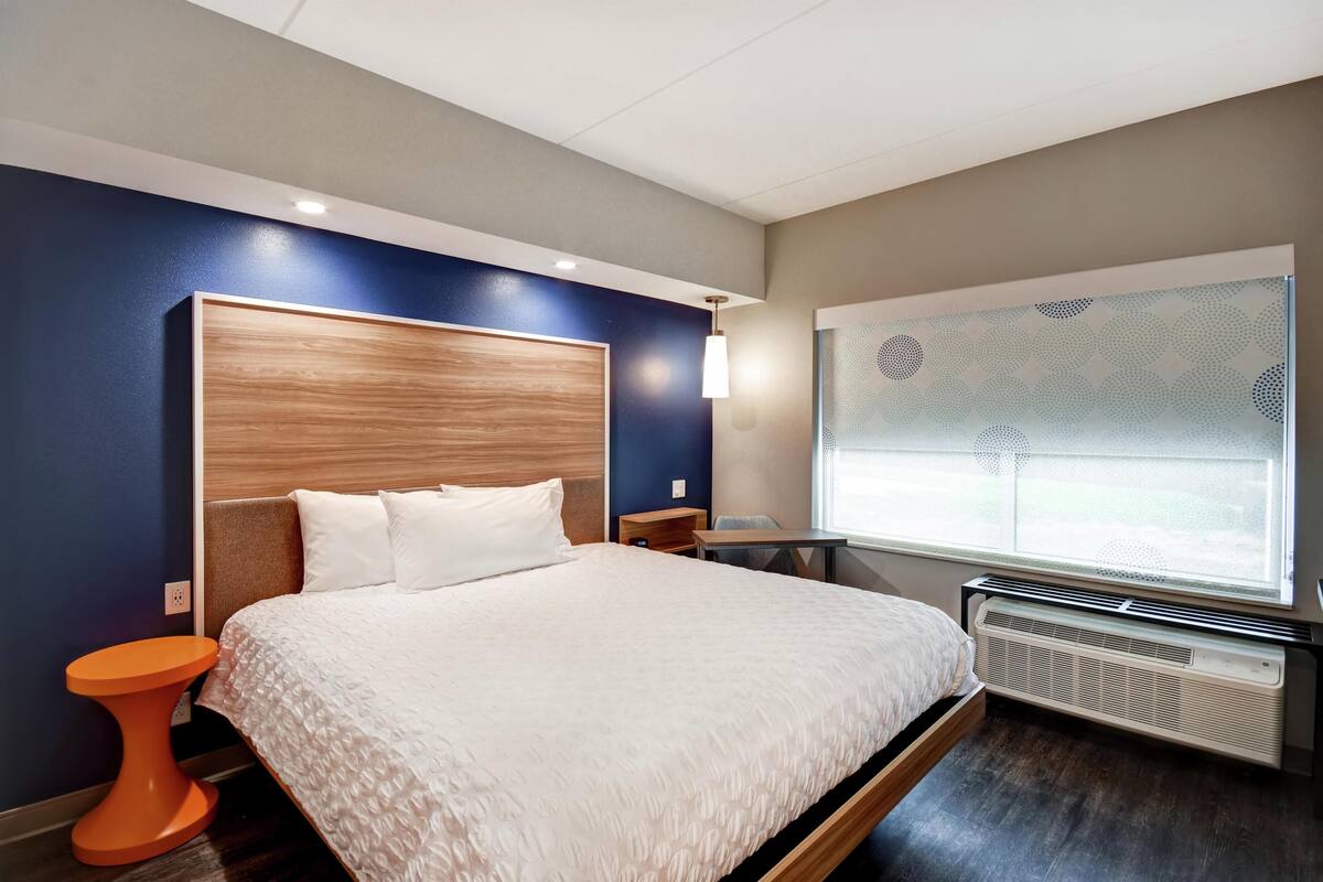 A modern hotel room with a large bed, a small table, a chair, a heater, and a window with a geometric pattern.