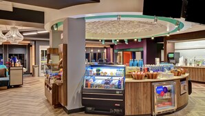 A modern hotel lobby with a central counter, a refrigerated display case, and a seating area with a sofa. - Tru by Hilton Raleigh Durham Airport (Morrisville)