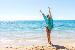 Una persona en la playa, de brazos alzada, vestida con una blusa azul y tanga roja.