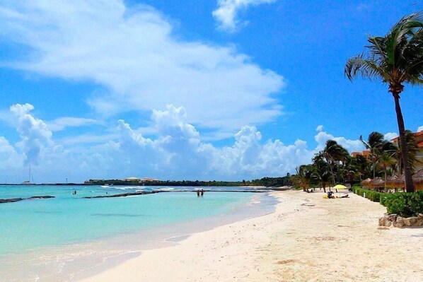 Una playa con agua turquesa cristalina, palmeras y algunas personas a lo lejos.