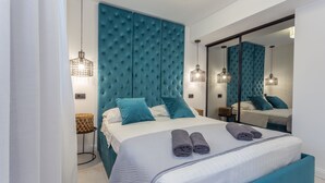 Ein modernes Schlafzimmer mit einem großen Bett, einem mit Türkis bezogenen Kopfteil und passenden Kissen. Zwei Nachttische mit Lampen, ein großer Spiegel und ein Fenster mit Vorhängen sind ebenfalls vorhanden.
