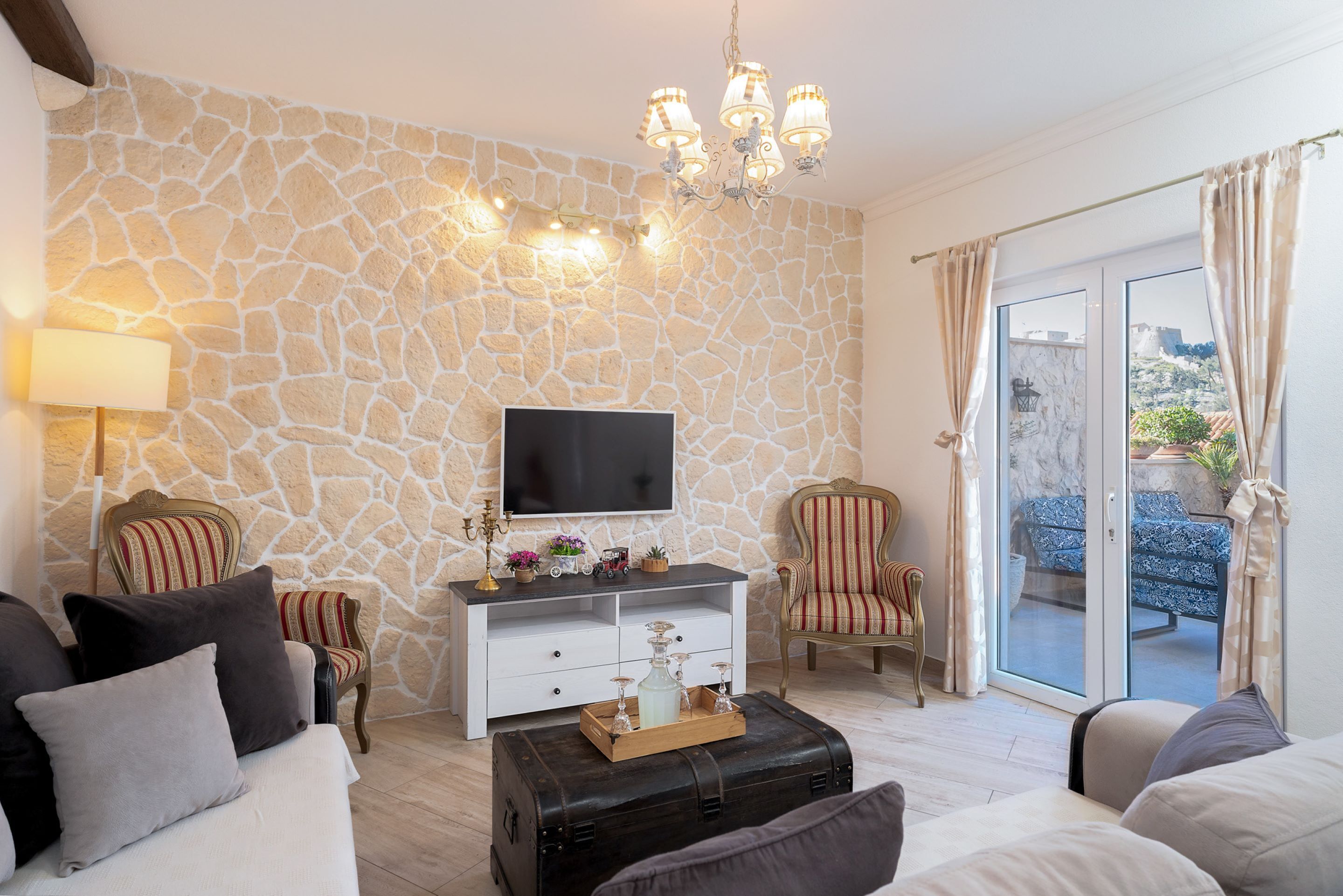 A living room with a stone wall, a flat-screen TV, a wooden coffee table with a tray, and two armchairs.