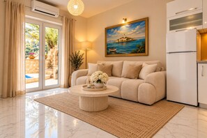 Un moderno salón con un sofá beige, una mesa de centro redonda con un jarrón, un cuadro con una escena costera y una zona de cocina con un refrigerador blanco.