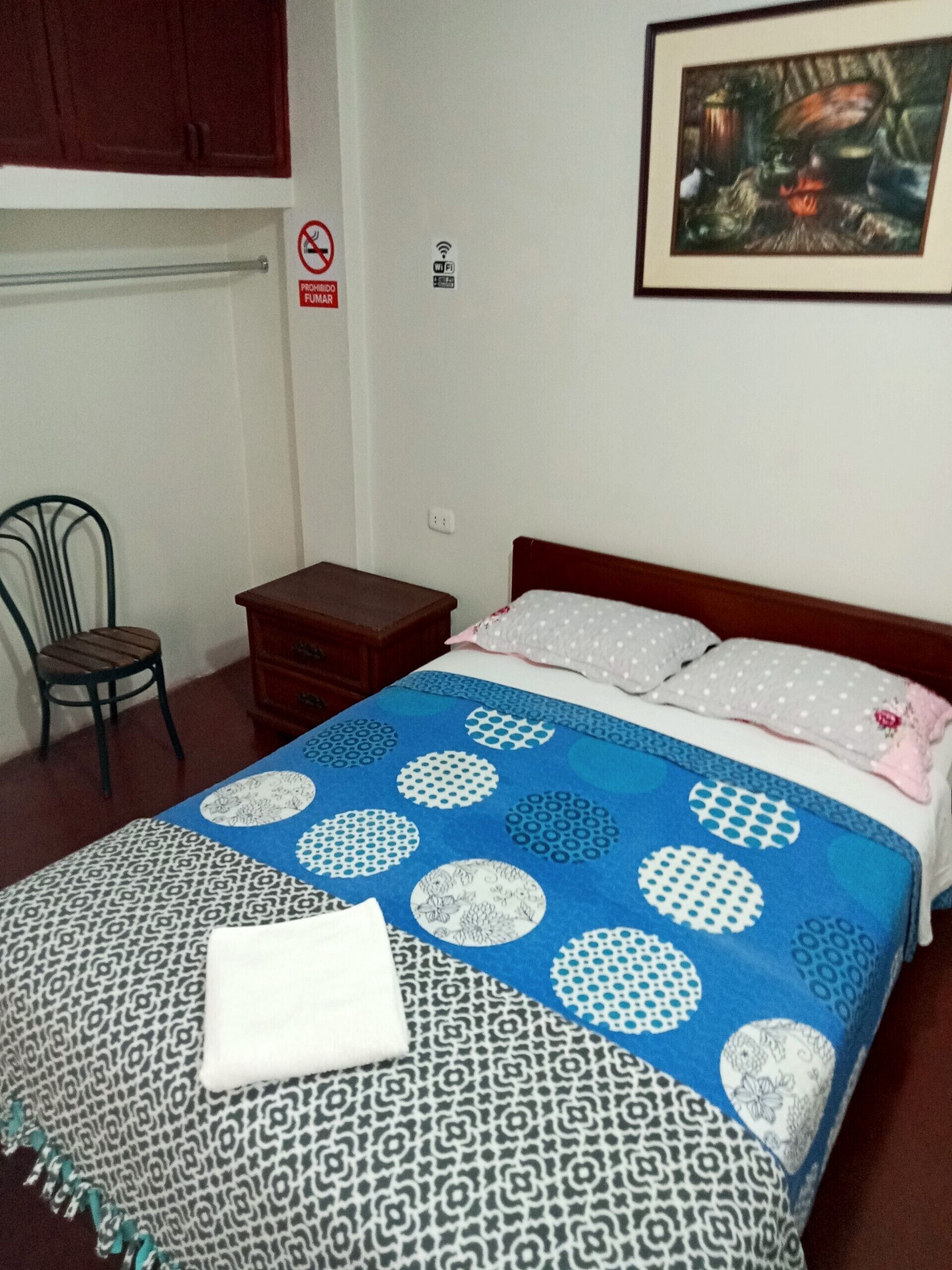 Un dormitorio con cama, una silla, una mesita de noche, un cuadro en la pared y un cartel de Wi-Fi.